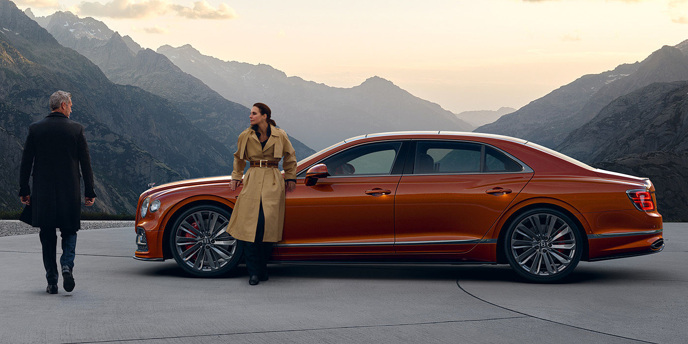 Bentley Baku Bentley Flying Spur Speed parked in Orange Flame coloured exterior parked, with mountainous background and two people in view.